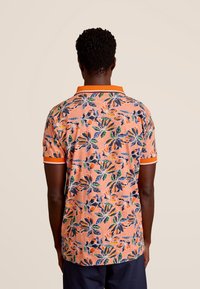 Polo shirt met korte mouwen op een koraalkleurige achtergrond, met veelkleurige bloemenpatronen, een oranje kraag en contrasterende mouwen.