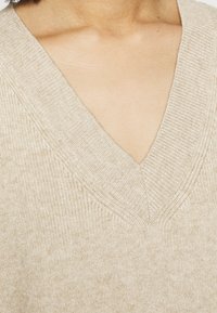 Beige V-neck neulepaita, joka on valmistettu pehmeästä, ribatusta neuloksesta. Ominaisuuksiin kuuluu syvä pääntie ja teksturoitu kuvio reunoilla.