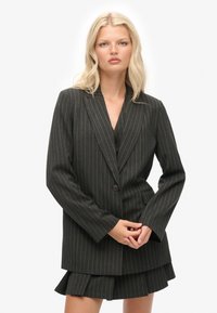 Sort blazer med striber, med spidse klaffer, enkelt knaplukning og lange ærmer. Bæres med en lagdelt nederdel, hvilket skaber et formelt look.
