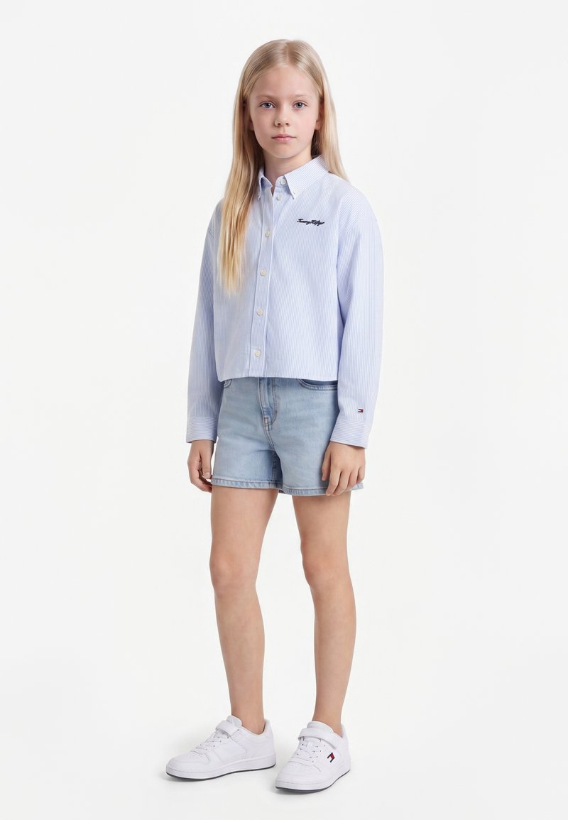 Fille blonde debout face à l'avant, portant une chemise bleu clair boutonnée, un short en jean et des baskets blanches sur un fond blanc.
