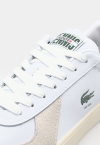 Baltos odinės sportinės bateliai su smėlio spalvos zomšo akcentais, žalia emblema ir baltos raišteliais. Yra guminis padas ir etiketė su užrašu "Lacoste Tennis."