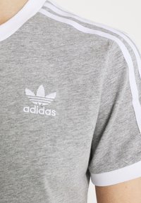 Pilkas marškinėliai su balta kraštine aplink apykaklę ir rankovių kraštus, kuriuose yra trys baltos juostelės pečių srityje ir siuvinėtas Adidas logotipas.