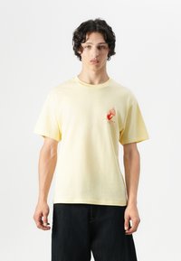 Camiseta de algodón amarilla con un pequeño diseño gráfico rojo en el pecho. Cuello redondo simple y mangas cortas. Combinada con jeans oscuros.