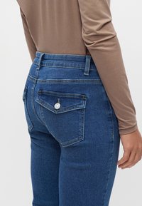 Personne portant un jean bleu taille haute avec un détail de poche arrière boutonnée et un haut beige à manches longues, debout devant un fond uni.