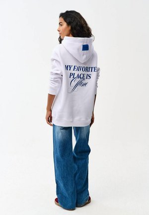 Kobieta z długimi ciemnymi włosami, odwrócona tyłem, ma na sobie biały hoodie z niebieskim napisem "MY FAVORITE PLACE IS Offline" i szerokie niebieskie dżinsy.