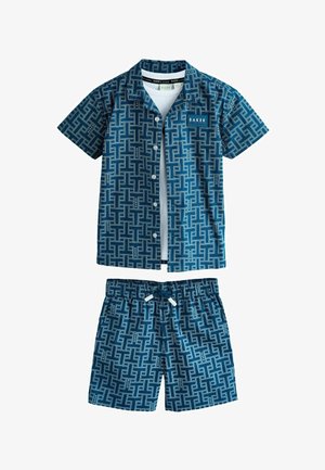 Modrá košeľa s krátkym rukávom na gombíky a zodpovedajúce krátke nohavice s geometrickým vzorom, pod ktorými je vrstvené biele tričko.