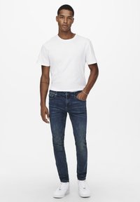 Fehér, rövid ujjú póló, sötétkék, slim fit farmernadrág, amelyen fakulás látható, és fehér sportcipő. Egyszerű dizájn, hétköznapi öltözet.