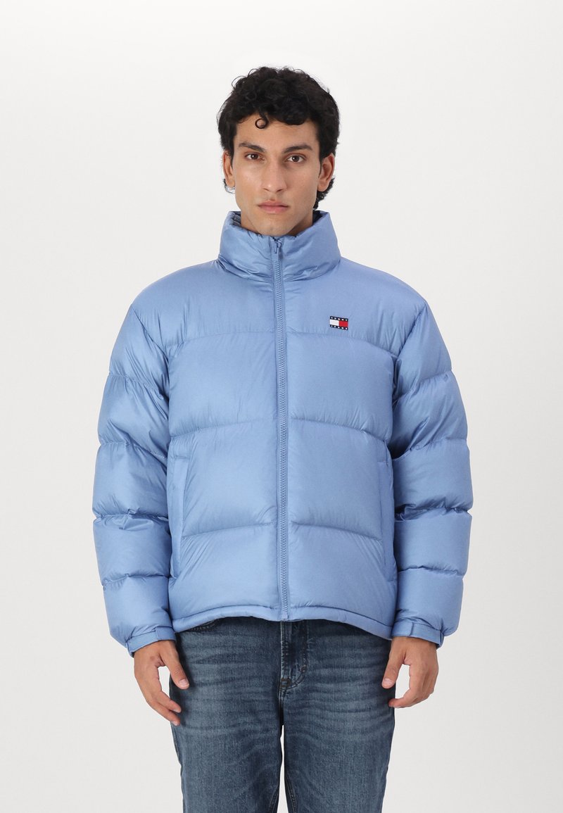 Hellblaue Steppjacke mit hohem Kragen, ausgestattet mit einem Reißverschluss und gesteppten Paneelen. Kleines Logo auf der Brust, kombiniert mit Jeans.