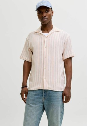 Férfi világos rózsaszín csíkos rövid ujjú, gombos ingben, kék farmerben, világoskék sapkában és fekete karkötővel, egyszínű világos háttér előtt áll.