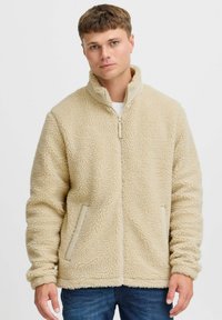 Beige fleece jacket with a high collar, zip closure, and side pockets. Soft texture with a slightly plush appearance.