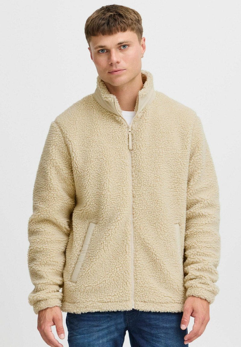 Beige fleece jacket with a high collar, zip closure, and side pockets. Soft texture with a slightly plush appearance.