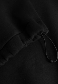 Felpa nera con cappuccio in tessuto, presenta un polsino arricciato con cordino e fermaglio, caratterizzata da una texture morbida e una finitura liscia.