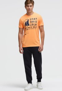 Orange cotton t-shirt featuring a graphic print with text and a sailboat design. Paired with dark navy sweatpants and white sneakers.