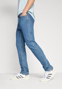 Jeans de mezclilla azul claro con piernas rectas, costuras laterales y costuras en contraste, combinados con zapatillas blancas y grises.