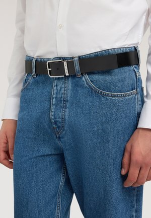 Man draagt een blauwe spijkerbroek en een wit overhemd met een zwarte leren riem die bij de taille is bevestigd met een zilveren gesp.