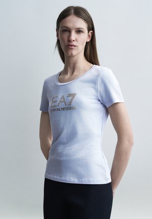 Žena nosi priliehavé svetlofialové tričko s logom "EA7 Emporio Armani" v zlatých cvokoch, stojaca pred jednofarebným sivým pozadím.