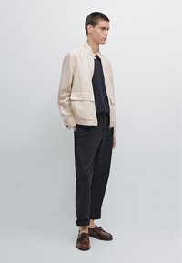 Beige bomberjack met twee voorzakken, gecombineerd met een donker shirt en losse, donkere broek. Bruine veterschoenen maken de outfit compleet.