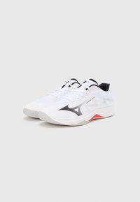 Chaussures de sport blanches avec une tige en mesh texturé, ornées d'accents noirs et rouges, un logo noir bien visible, et une semelle amortie.