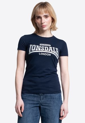Mujer con cabello corto y rubio, vestida con una camiseta azul marino de Lonsdale London y jeans azules, de pie frente a un fondo blanco liso.