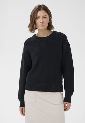 Femme aux cheveux bruns mi-longs portant un pull en maille noir et une jupe beige, debout devant un fond clair uni.