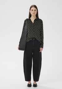 Fekete pöttyös blúz galléros nyakkivágással, magas derekú fekete farmerral viselve. A modell fekete válltáskát hord, és fekete lapos cipőt visel.