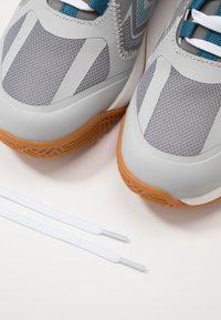 Grijze sportschoenen hebben een gestructureerde mesh bovenkant, teal accenten en een rubberen zool. Twee witte veters met plastic tips zijn eronder gepositioneerd.