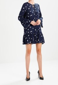 Laivansinisestä mekosta, jossa on valkoisia lintukuvioita, on kellohihat ja röyhelöhelma. Mekko sisältää nauhakujaan ja on valmistettu kevyestä kankaasta.