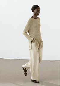 Maglione beige lavorato a maglia con maniche lunghe, aperture laterali, abbinato a pantaloni a gamba larga beige chiaro e ciabatte nere. Tessuto morbido e texture.