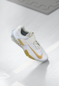 Zapatilla deportiva blanca con un distintivo swoosh dorado de Nike, parte superior de malla, collar acolchado y un cierre con correa singular, además de una suela translúcida.