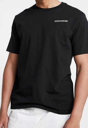 Black short-sleeve crew neck t-shirt with small white "Converse" logo on the left chest, worn with white pants.