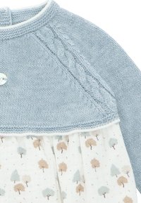 Top de suéter azul de punto con patrón de trenzas, detalle de botones y falda blanca con estampado de árboles en beige y azul claro.