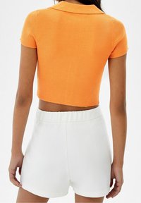 Top corto a maniche corte arancione a coste abbinato a pantaloni corti bianchi a vita alta, con una cintura elastica e una texture liscia.
