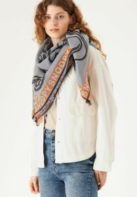 Grauer Schal mit schwarzem Snoopy-Motiv, umrandet von orangefarbigem Text. Weiche Textur mit fransigen Kanten. Wird um den Hals drapiert getragen.