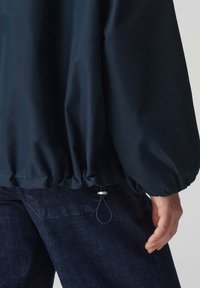 Marineblauwe windjack met een elastische zoom, met een gladde buitenkant. Verstelbare trekkoord met metalen accent in de taille. Donkerdenim broek zichtbaar.