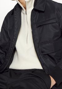 Svart bomberjacka med en slät tyg, med dragkedja framtill, sidofickor och ribbade muddar, bärs över en vit ribbad stickad tröja.