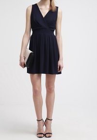 Modelka ve tmavě modrých šatech bez rukávů s přiléhavým korzetem a rozšířenou sukní, držící černobílou kabelku, spárovanou s černými sandály na podpatku.