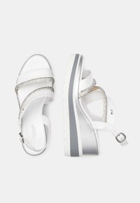 Sandalias de cuña blancas con acentos plateados, que presentan correas entrelazadas con detalle metálico, hebilla ajustable en el tobillo y suela texturizada.
