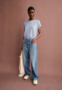 Light blue short-sleeve t-shirt, loose-fit light denim jeans, holding a cream-colored cardigan, and gray sneakers with green accents.
