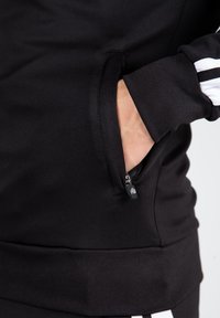 Sweatshirt noir avec une poche zippée, des poignets côtelés et un ourlet. Présente des rayures blanches sur les manches. Texture lisse.