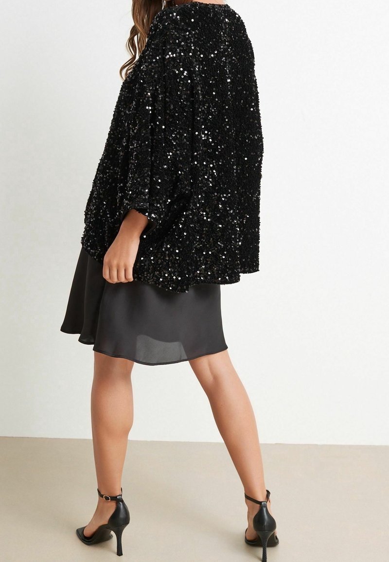 Femme portant une veste noire ample à paillettes sur une jupe en satin noire longueur genou et des talons aiguilles noirs à bride cheville, debout avec une jambe en avant.