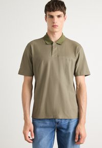 Olivgrünes Poloshirt aus glattem Baumwollstoff, mit Kragen, kurzen Ärmeln und einem Logo auf der Brust. Getragen mit blauen Jeans.