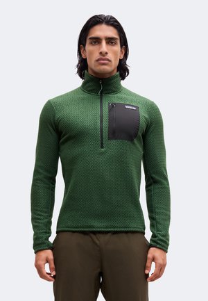 Jeune homme aux cheveux foncés porte un pull vert texturé à manches longues avec une poche poitrine zippée noire et un pantalon marron.