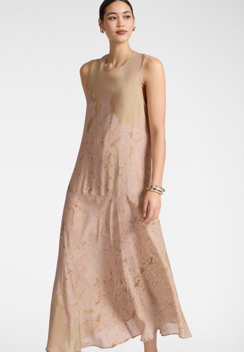 Mujer con vestido largo sin mangas beige-rosa con estampado de cachemira, pendientes de aro dorados y pulseras apiladas, de pie frente a un fondo liso.