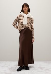 Bruine geruite blouse met lange mouwen, gecombineerd met een witte gebreide sjaal, en een bruine satijnen maxi rok en zwarte chunky schoenen. Soepele texturen en gelaagd ontwerp.