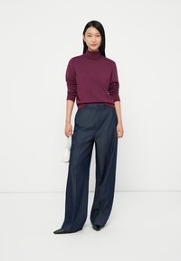 Pulôver de gola alta bordeaux combinado com calças de ganga soltas de um azul escuro. O modelo segura uma pequena bolsa branca; sapatos pretos com bico fino completam o visual.