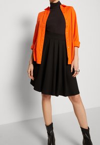 Camicia arancione con bottone, indossata sopra una gonna nera a pieghe, lunga fino al ginocchio. L'insieme è abbinato a stivaletti neri alla caviglia.