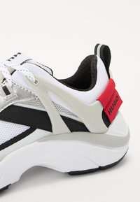 Baskets blanches avec accents argentés et noirs, comportant une tige en maille texturée, une semelle épaisse blanche et une bride rouge avec logo au talon.