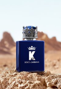 Flacon de parfum Dolce & Gabbana K bleu avec bouchon argenté en forme de couronne sur une surface rocheuse, montagnes désertiques floues en arrière-plan.