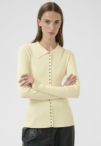 Młoda kobieta z długimi brązowymi włosami, ubrana w kremowy sweter z guzikami w karbowanym wzorze i czarne perforowane spodnie, z założonymi rękami, na neutralnym tle.