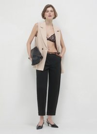 Blazer beige senza maniche sopra pantaloni neri a vita alta, bralette in pizzo nero, che tiene una clutch nera, con tacchi neri a punta.